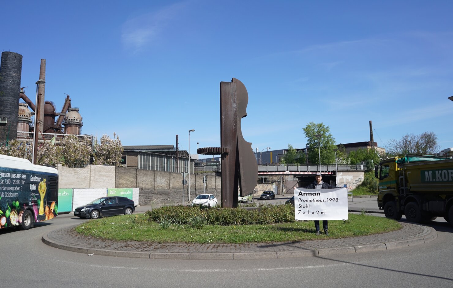 Matthieu Martin holding hand-painted road-sign-style label next to roundabout sculpture, Kreisverkehr project, 2026