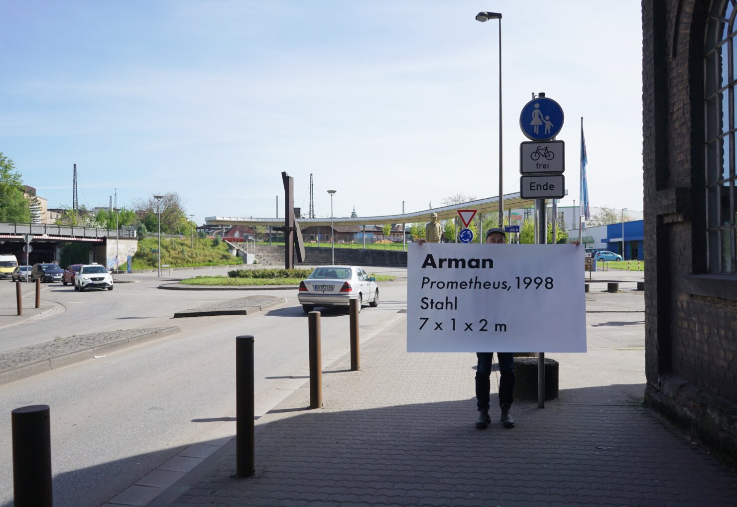 Matthieu Martin holding hand-painted road-sign-style label next to roundabout sculpture, Kreisverkehr project, 2026